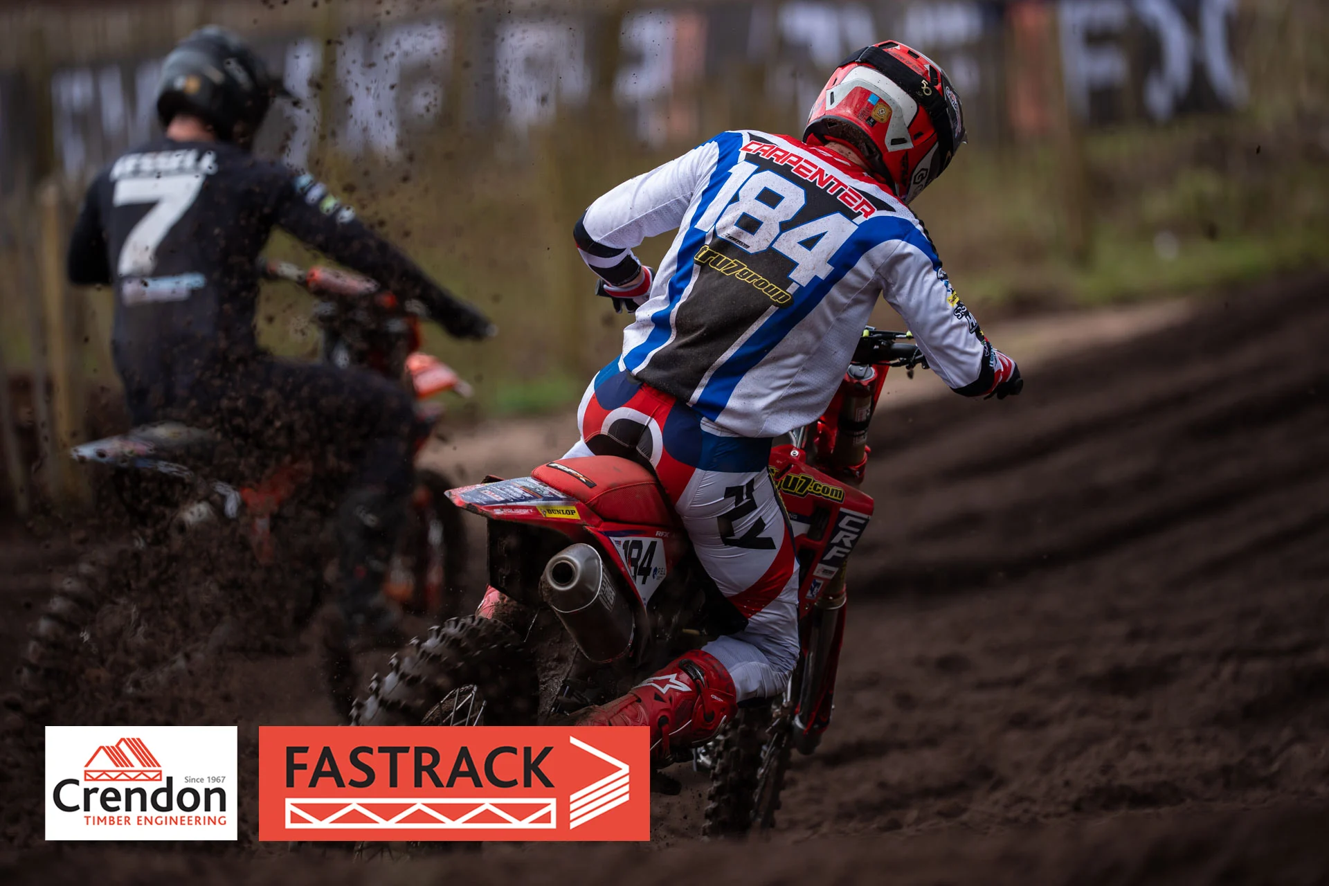 View of the back of a biker wearing red, white and blue on a dirt track with a rider wearing black in the back left corner. 