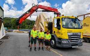 Three workers in front of a yellow lorry holding roof trusses with a crane attached