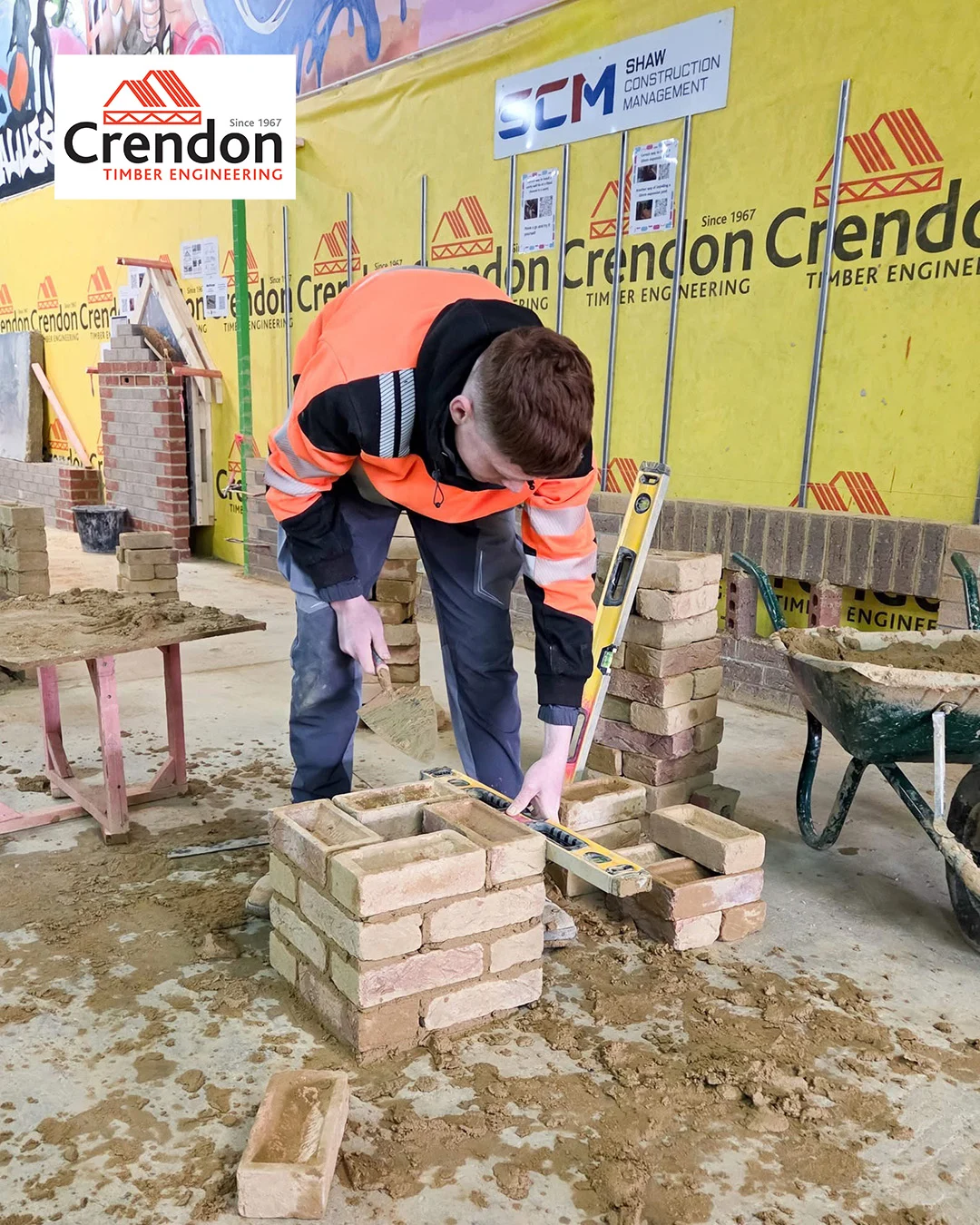 Worker in orange hi-vis jacket using a spirit level on a small brick build in a workshop with Crendon signage.